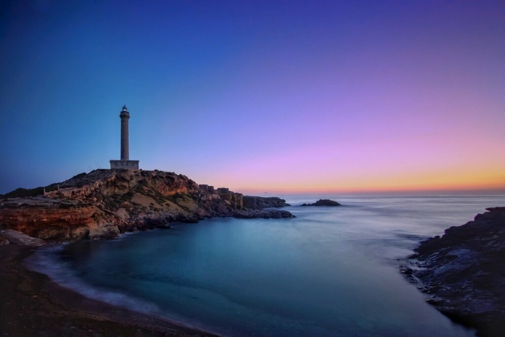 Leuchtturm auf Insel vor Festland auf blauem Wasser vor blauem Hintergrund, letztes Abendrot am Horizont