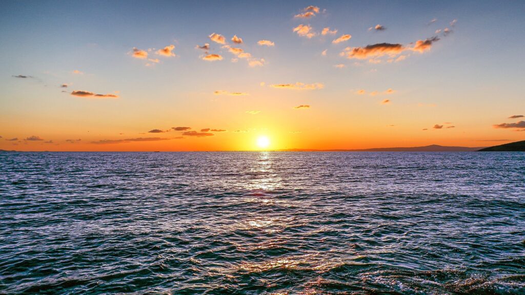 Sonne versinkt orange über dem blauen Meer, am Bildrand ist eine Insel erahnbar